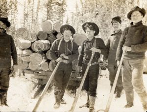 Sepia photograph of a logging camp: five men posing in a snowy environment. Warm clothing, high boots. Four men with hats, one with a cap. Each holds a pulp hook. behind: large pile of logs on a sleigh. Background: snow-covered trees. Atmosphere of hard labour in the winter.