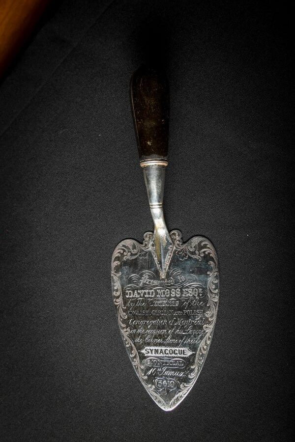 An inscribed metal trowel on a black surface. The inscription reads Presented to David Moss Esquire, by the trustees of the English, German and Polish Congregation of Montreal on the occasion of this Laying of the Corner Stone of their Synagogue. Montreal, 40th Tammuz, 5619.