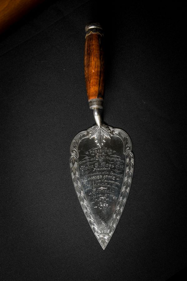 An inscribed metal trowel on a black surface. The inscription reads Presented by the German and Polish Congregation to John E. Moss Esquire, President, on the occasion of his laying the corner stone of the new synagogue on McGill College Ave. Montreal. Heshvan, 12th, 5656. October 21st, 1885.