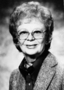 Black and white head and shoulders photographic portrait of a smiling middle aged woman with curly hair and glasses wearing a woolen jacket over dark buttoned shirt and neck scarf.