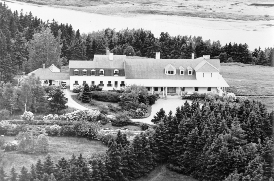 Black and white aerial photograph of the Reford Gardens. On the right side of the photograph is a clump of spruce trees, and on the left is a long flower bed. In the upper part of the photograph is the Estevan Lodge, a long white wooden residence with a dark roof. At the top of the picture, a river crosses from left to right.