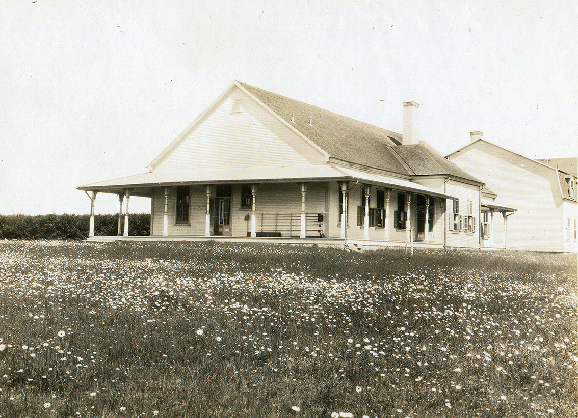 Black and white photograph of the north and west facades of Estevan lodge. The exterior of the building is in wood and the gable roof is made of cedar shingles. The windows are equipped with shutters. The fishing camp is equipped with a generous verandah on which is placed rack for fishing rods. At the back of the main building are outbuildings occupied by the owner's servants. The two-storey building is slightly set back to the west in relation to the main building. The building is surrounded by a flowering meadow. We can see the huge cedar hedge that occupies the front space of the fishing camp. In spite of its size, the fishing camp is rather sober, only the columns of the verandah roof underline the Victorian style of the building's architecture.