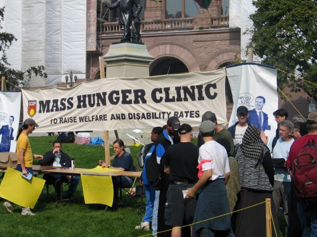 A large banner with the words Mass Hunger Clinic to Raise Welfare and Disability Rates in front of the Ontario legislature. Two people are seated at an information table. Other people are lined up or engaged in conversation.