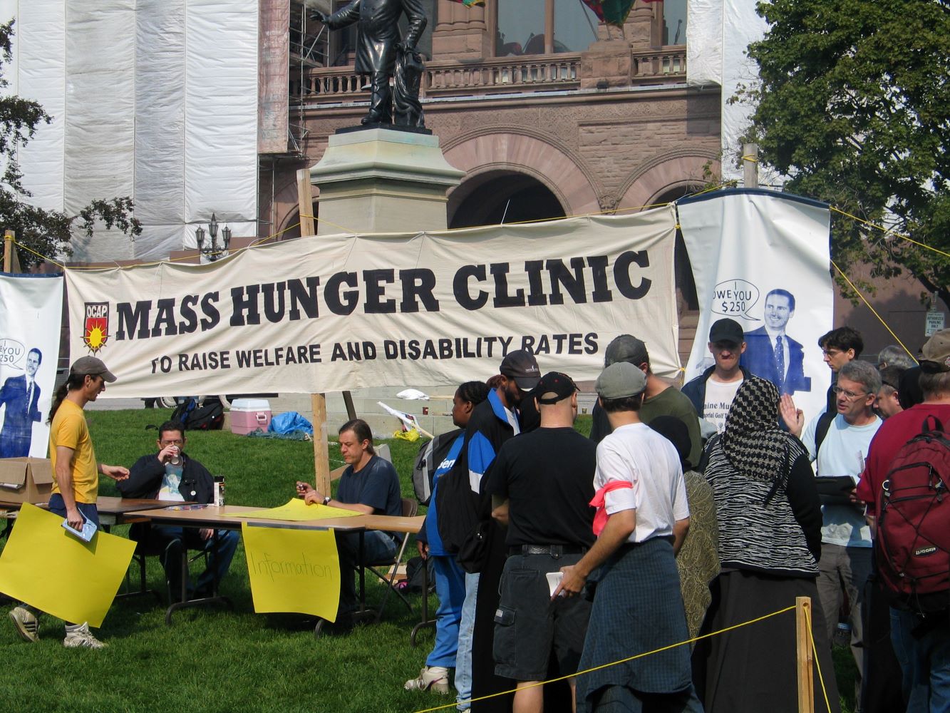 A large banner with the words Mass Hunger Clinic to Raise Welfare and Disability Rates in front of the Ontario legislature. Two people are seated at an information table. Other people are lined up or engaged in conversation.