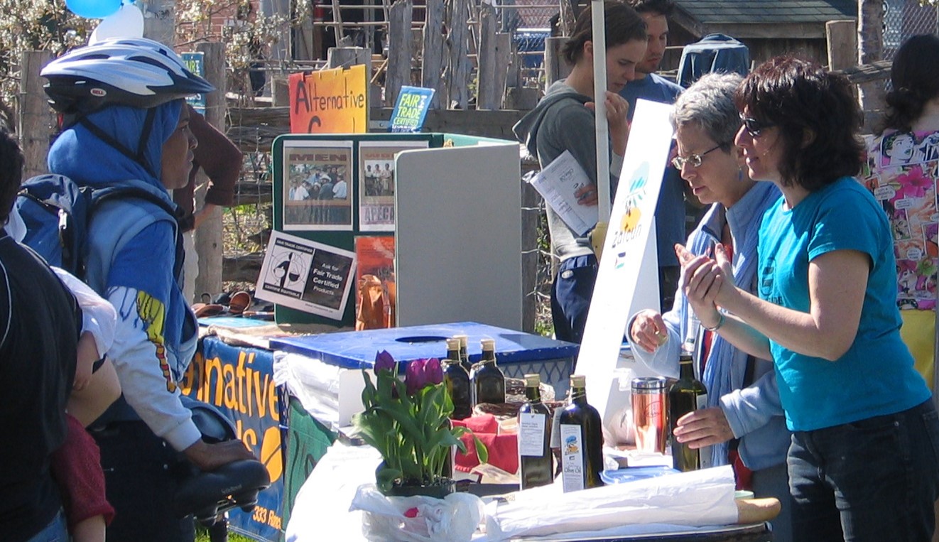 A woman standing behind an outdoor vendor table with bottles of Palestinian olive oil, talking to another woman.