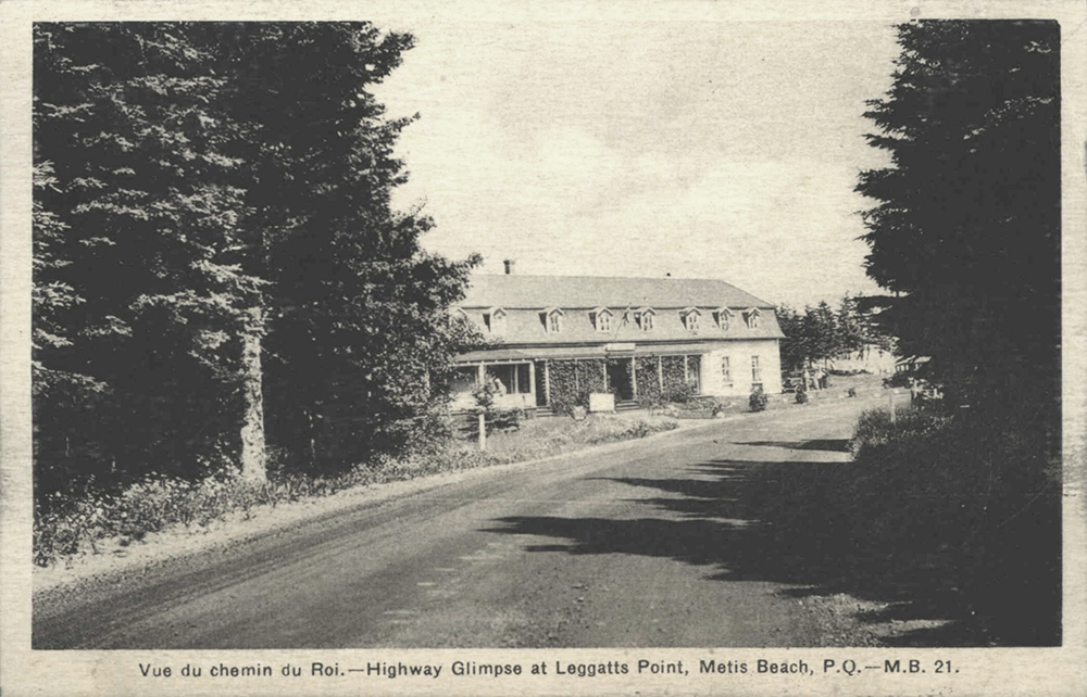 Postcard with a black and white photograph of the Killiecrankie Inn. In the foreground, a dirt road bordered by a spruce forest. Between the spruce trees on the left side of the road appears Killiecrankie Inn. The inn is a long 2-storey building made of wood. The facade has a veranda invaded by a vine.