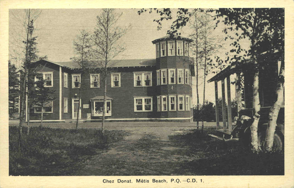 Black and white photograph of the Chez Donat hotel. The two-storey hotel is located on the side of the road. A three-storey turret forms the right corner of the building. Each window is decorated with white curtains. The photograph was taken from the courtyard entrance in front of the hotel.