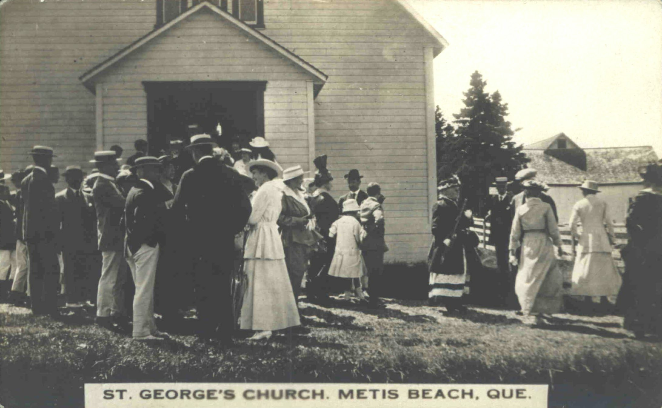 Black and white photograph of men and women gathered in front of a wooden church at the end of mass. The men are, for the most part, dressed in a dark jacket, a white pentalon and a flat braided hat decorated with a ribbon. The women wear a long white dress and a flat braided hat. In the background, a farm building can be seen behind a clump of mature spruce trees.