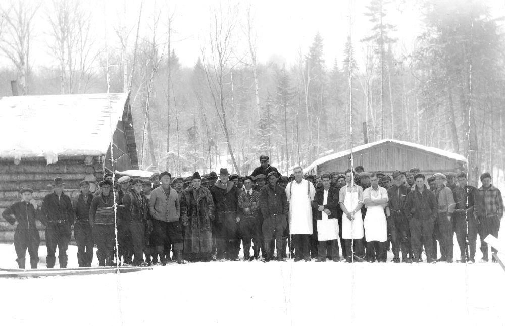 Black and white photograph of a logging camp: large group of men in front of two snow-covered wooden buildings. Men in warm clothing, some with white aprons (cooks). Background: forest, bare trees, overcast sky. Atmosphere of an isolated working community.