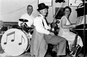 Black and white photograph of three smiling seated adult musicians, two men and a woman, with instruments.