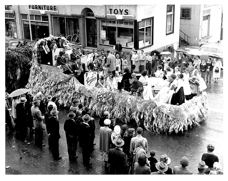 Black and white photograph looking down at a paved street lined with people watching a trailer, decorated as a parade float the sides of which are trimmed with cedar boughs. At the front of the trailer is seated a musical band of about 15 persons being conducted by a man standing facing them, with his back to about 8 people seated at the back of the float.