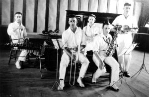 Black and white photograph of five short haired young male musicians with instruments dressed identically in open collared white shirts, white trousers and white shoes.