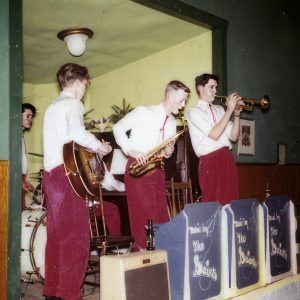Colour photograph of five clean shaven young musicians with short haircuts wearing white long-sleeved shirts, bolo ties and red trousers standing on a stage playing trumpet, saxophone, acoustic guitar, drums and piano.