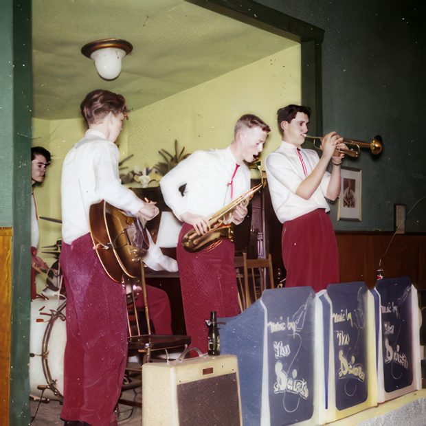 Colour photograph of five clean shaven young musicians with short haircuts wearing white long-sleeved shirts, bolo ties and red trousers standing on a stage playing trumpet, saxophone, acoustic guitar, drums and piano.