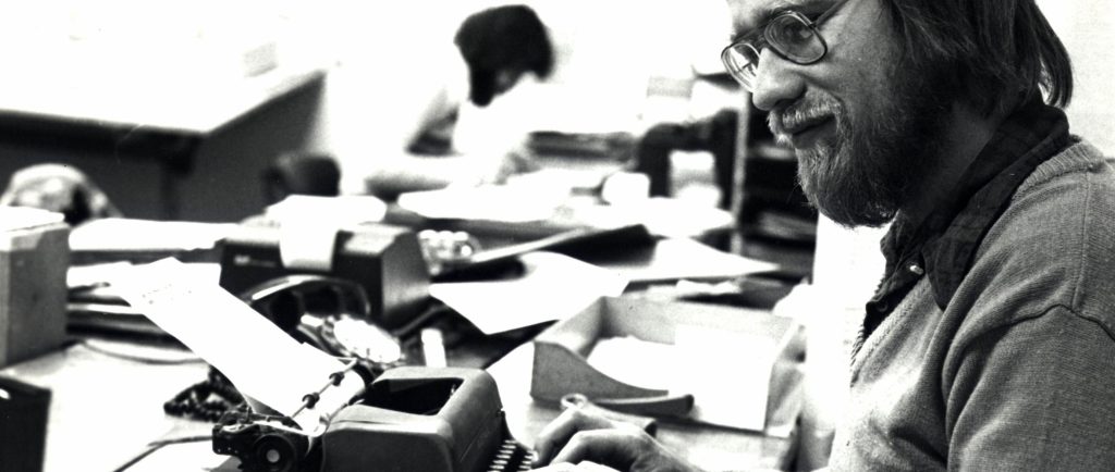 A bearded man typing on a small manual typewriter which has a sheet of paper fed onto the roller.