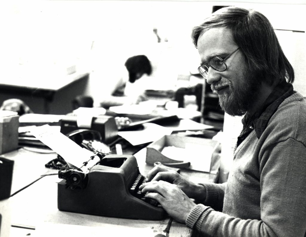 A bearded man typing on a small manual typewriter which has a sheet of paper fed onto the roller.