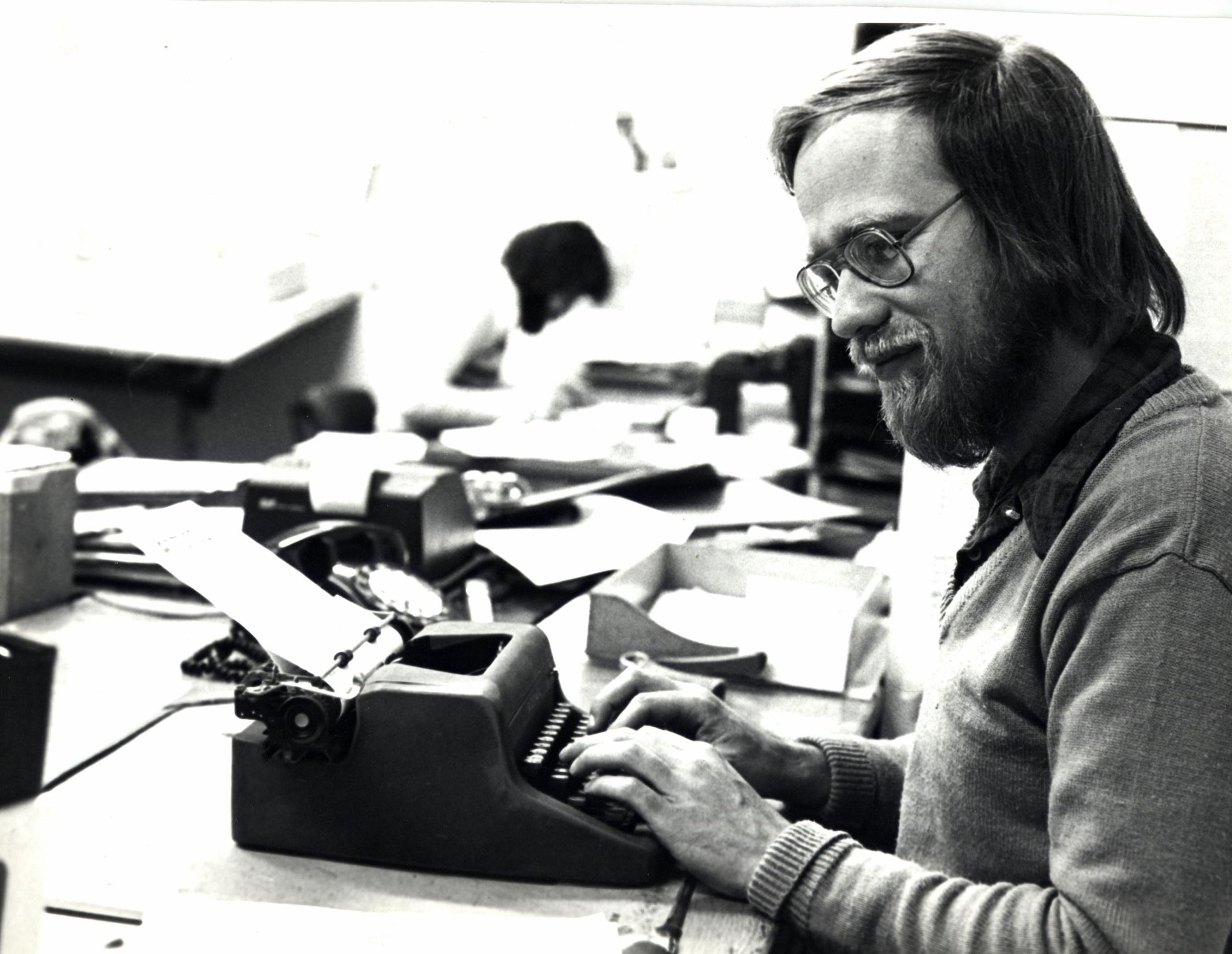 A bearded man typing on a small manual typewriter which has a sheet of paper fed onto the roller.