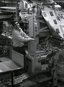 A press operator standing at the controls of a large web offset press smiles at the camera while a continuous sheet of freshly printed newspaper rolls off the press. At the bottom left, individual copies of the same newspaper are visible after they have been cut and folded.
