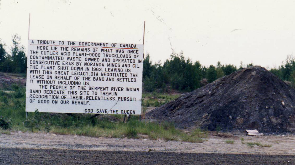 A sign on the side of the highway, with a dark pile of toxic waste beside it on the right. The sign says: A Tribute to the Government of Canada. Here lie the remains of what was once the Cutler Acid Plant - 9000 truckloads of contaminated waste. Owned and operated in consecutive eras by Noranda Mines and CIL, the plant shut down in 1963, leaving us with this great legacy. DIA negotiated the lease on behalf of the Band and settled it without including us. The People of Serpent River Indian Ban dedicate this site to them, in recognition of their relentless pursuit of good on our behalf. God Save the Queen.