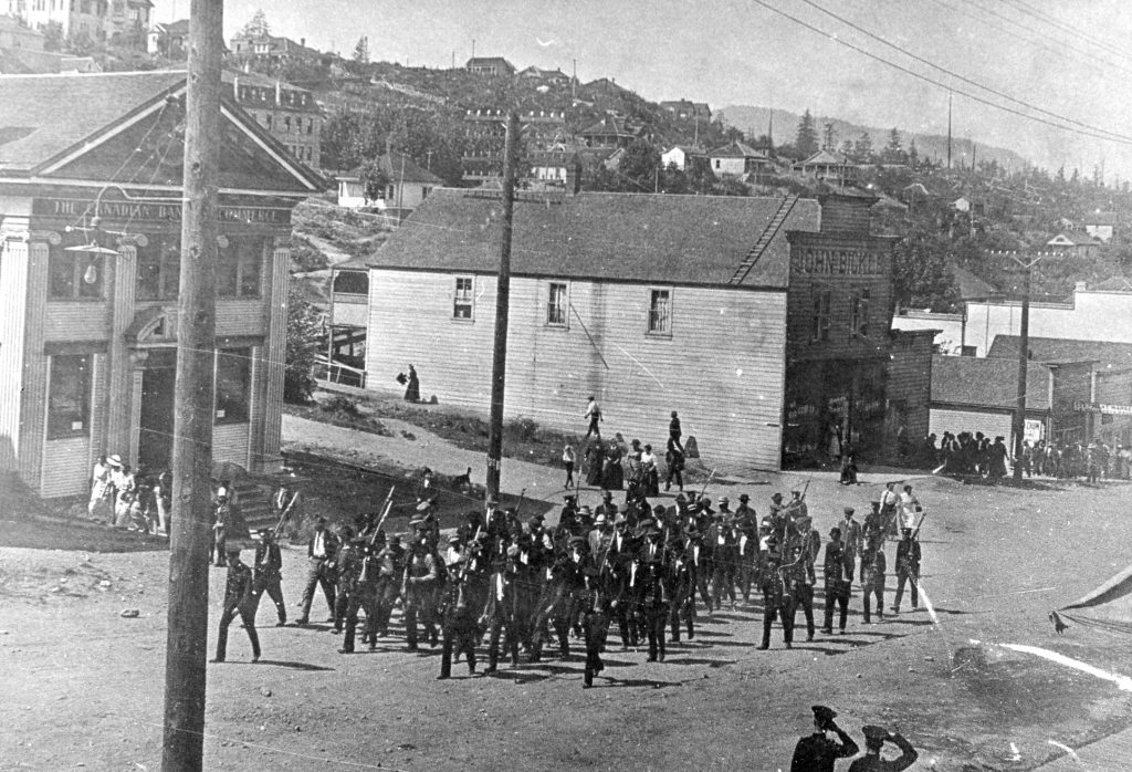 Black and white photograph of an unpaved town street along which march armed soldiers flanking a group civilians.