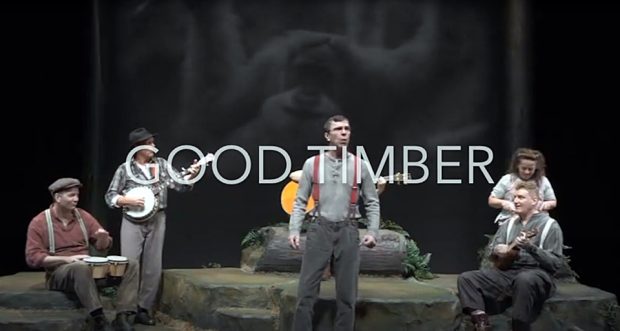 Colour photograph of five musicians on stage. The lead singer is dressed in work clothes and suspenders.