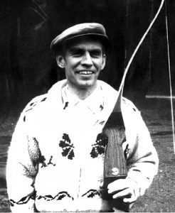 Black and white photograph of a middle aged craggy featured man wearing a cap and heavy woollen cardigan holding a wooden archery bow in his left hand.