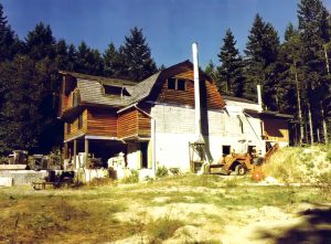 Colour photograph of an extensive barn-style three-story building in the shape of a cross, the centre portion of which is constructed of cement and cement block with the ends and upper stories made of wood.