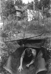 Black and white photograph of man crawling out of an underground passage. A three-story barn-like structure is in the background about 100 meters behind where the man emerges.