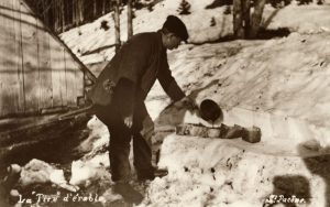 Sepia photograph, making maple taffy (S. Pacôme): a man in winter clothing pours maple taffy into bark containers on the snow. Background: snow-covered woods, part of a wooden structure on the left.