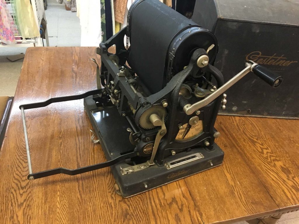 A black Gestetner machine on a wooden table. Its internal workings are visible from the outside: rollers, gears, dials and a crank. Behind it is a black case with the the word Gestetner in a script font written across it.