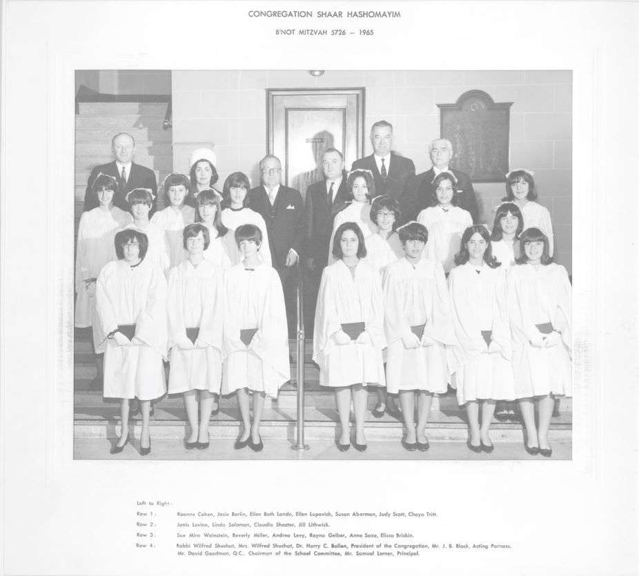 A group of adolescent girls in white graduation robes posing with teachers behind them.
