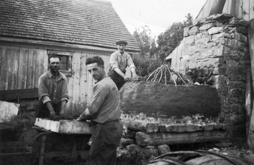 Black and white photograph (1930s): three men building an outdoor bread oven. Background: branches covered with earth or mortar. Behind: wooden building with a shingle roof. Scene of artisanal construction of an outdoor bread oven.