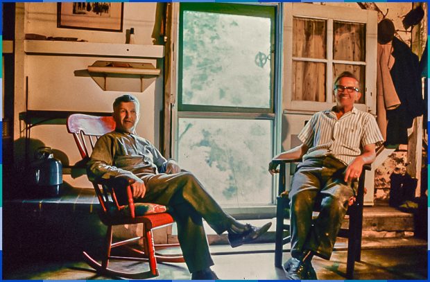 Two men, each sitting on a chair, in their home.
