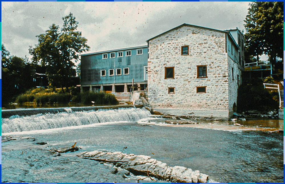 A colour photograph showing the south side of the Légaré mill. We can see the dike, the stone mill and the sawmill, which is connected perpendicularly.