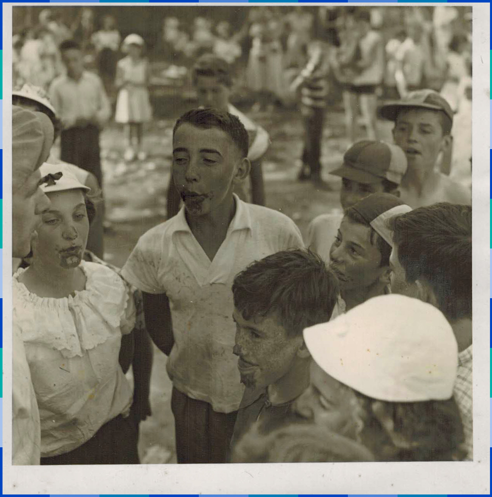 A black and white photograph. In the foreground, around ten muddy children who look to be about twelveyears old appear to be whistling. In the background, we can see around twenty other young people.