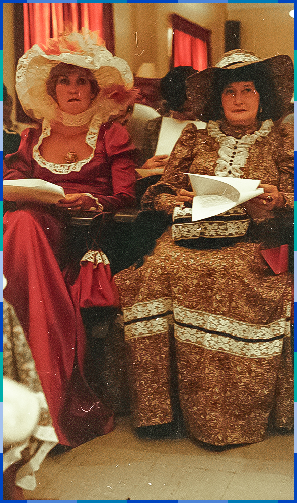 A colour photograph of two women wearing long colourful dresses and extravagant hats. They are sitting and attentively following the deliberations of the historical council. Behind them, we can see spectators.