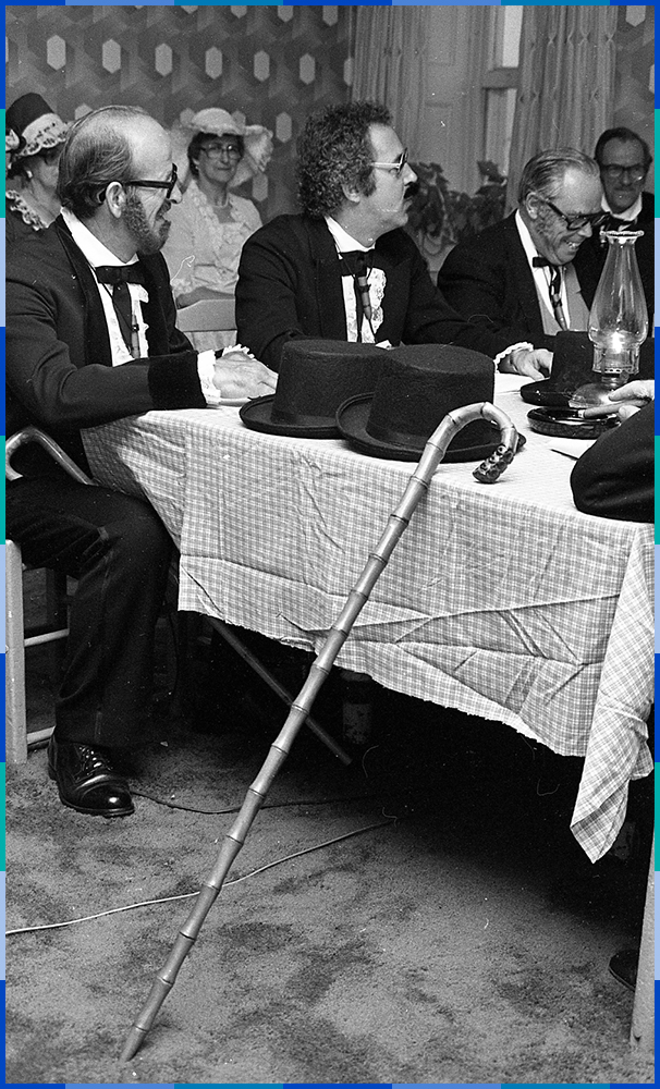 A black and white photograph of three councillors sitting at the corner of a table laughing. Sitting on the table are top hats, canes and an oil lamp.