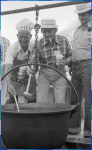 A black and white photograph of three men smiling as they watch a large pot. Two of them are holding ladles. The oldest of the three is wearing a hat that has the word “chef” on it, while the two others are wearing Saint-Eustache Knights of Columbus hats.