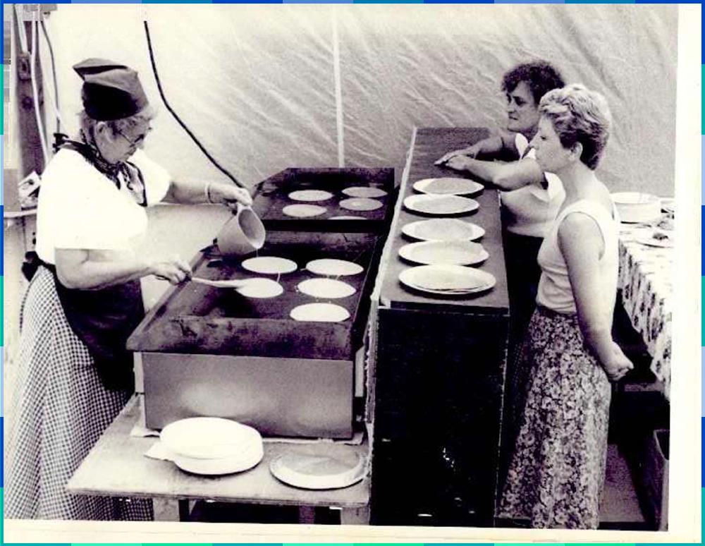 A black and white photograph of two women behind a counter watching a cook who is pouring pancake batter onto a hotplate. There are several plates full of pancakes on the counter.