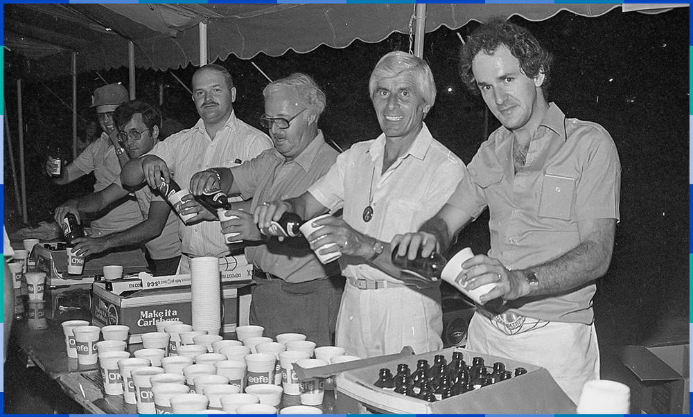 A black and white photograph of six men busy opening bottles of beer to pour it into small paper cups. In front of them is a table with dozens of cups and several cases of beer.