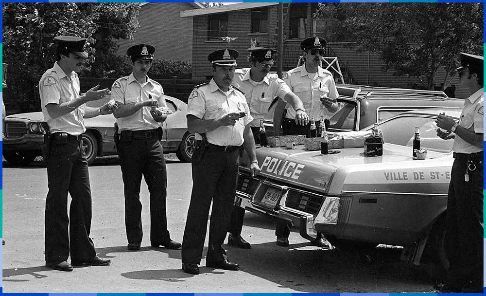 A black and white photograph of six police officers gathered around a patrol car. They are eating doughnuts and drinking Pepsi from glass bottles.