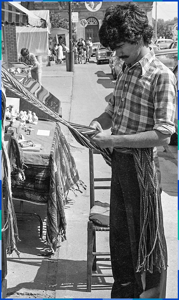 A black and white photograph of a craftsman making an arrow sash in front of his booth on rue Saint-Eustache. He is wearing one himself around his waist. Behind him are many people stopping at other booths. The Saint-Eustache Catholic church can be seen in the background.
