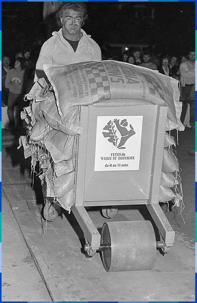 A black and white photograph of a man lifting a wheelbarrow containing more than ten sacks of flour. We can tell from his tensed expression that it is very heavy. A large crowd can be seen behind him.