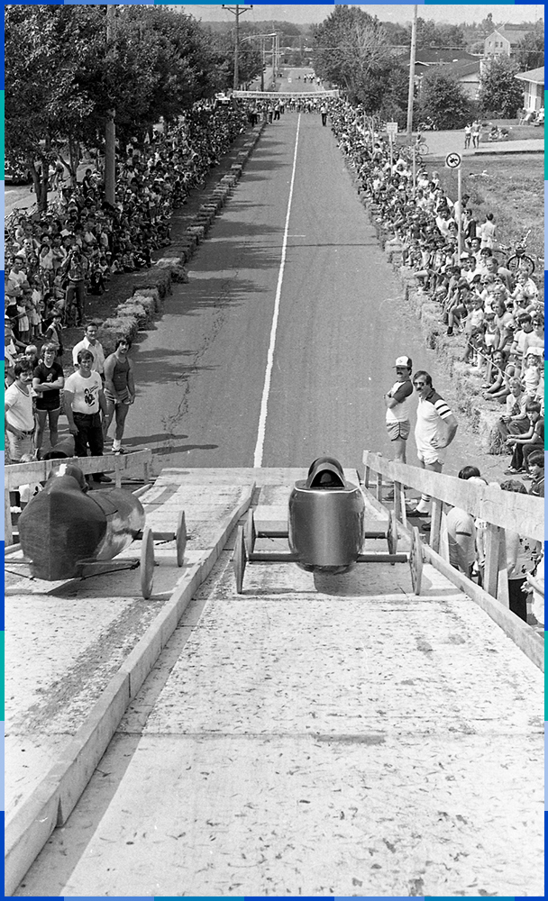 The photo is taken at the top of the soapbox ramp. A small vehicle is entering each of the two sections of the ramp. More than a hundred people are gathered on either side of the road, up to the finish line. The spectators are separated from the route by bales of hay.