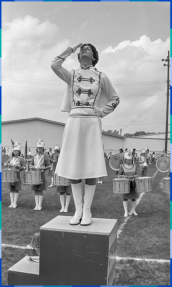 A black and white photograph. In the foreground, a woman standing on a podium is waving. Behind her, around ten other women are holding drums. They are all dressed in uniform that include boots and a white hat. They are on a grassy field.