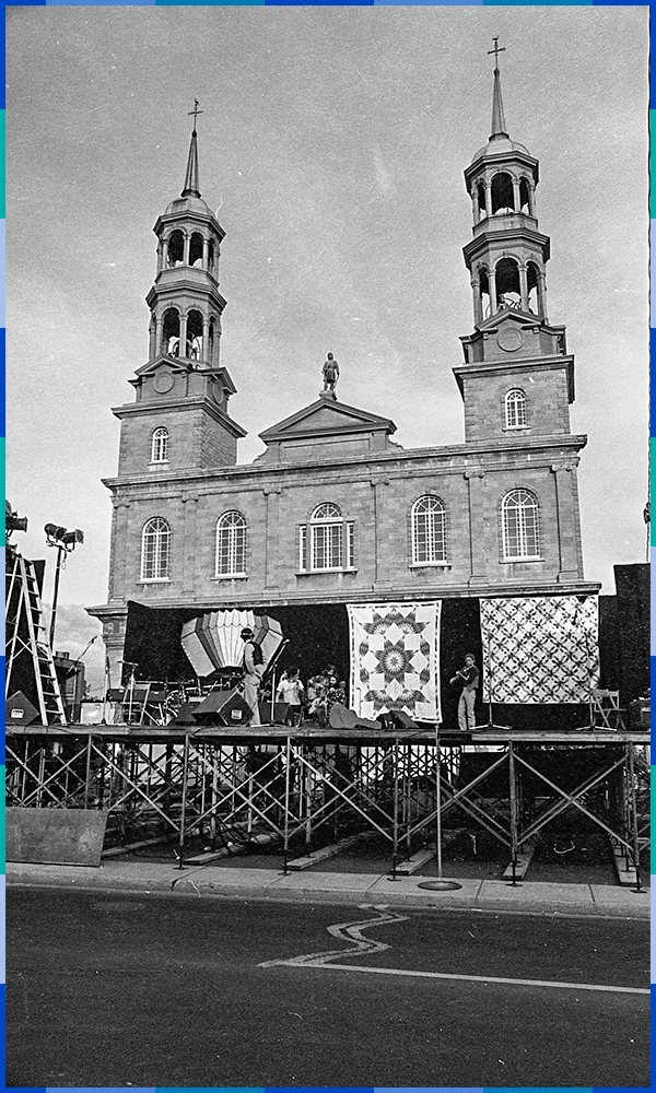 The black and white photograph shows a stage right in front of the Saint-Eustache church. On the stage, a group of musicians is rehearsing before the beginning of the Fêtes. The stage is decorated with large pieces of patterned fabric.