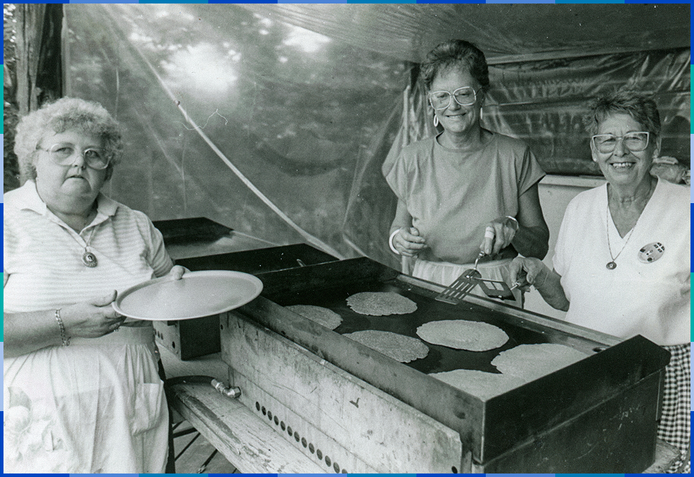 Two female cooks are making pancakes on a hotplate while a woman waits, holding a plate.