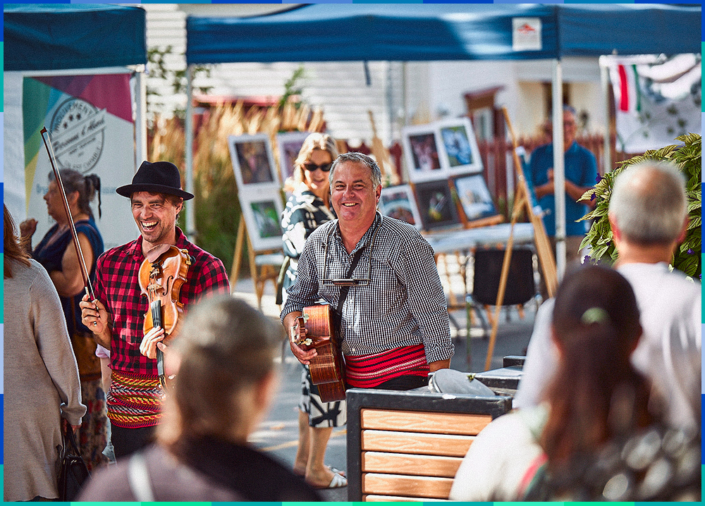 A photograph of two smiling men wearing plaid shirts and arrow sashes. One is holding a fiddle and a bow, while the other is holding a guitar. They are strolling among the booths and festival-goers.
