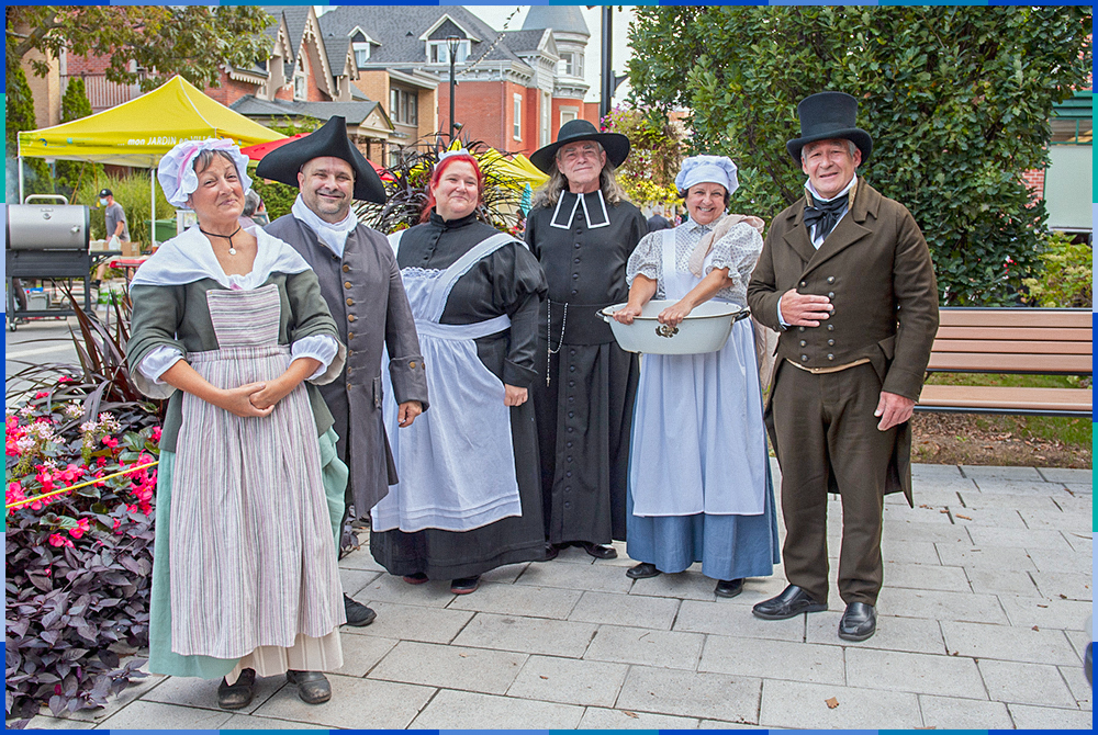 Six people, three men and three women, pose in Old Saint-Eustache. They are dressed in period costumes. Behind them we can see colourful booths.