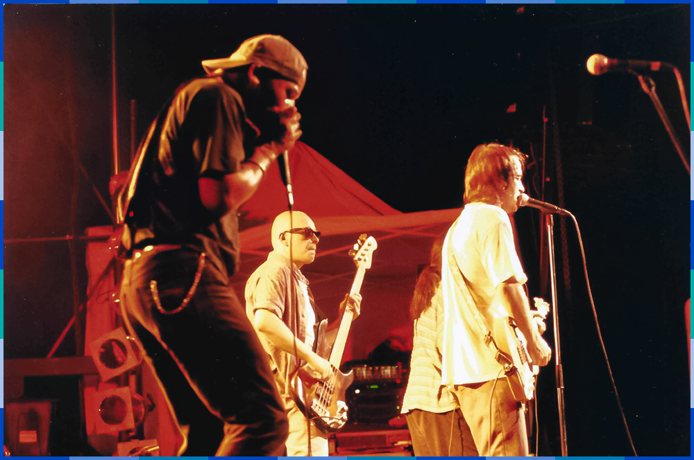 A colour photograph of the four members of the group Les Colocs, seen from the side, on a stage. Two of them appear to be singing into a microphone and two are playing the guitar.